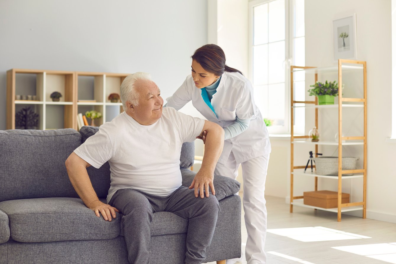 Nurse Helping Senior Man to Stand up from Sofa in Modern Hospital or Geriatric Rehabilitation Center